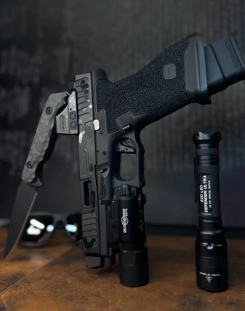 A stippled and milled pistol with a weapon light, beside a knife and a flashlight